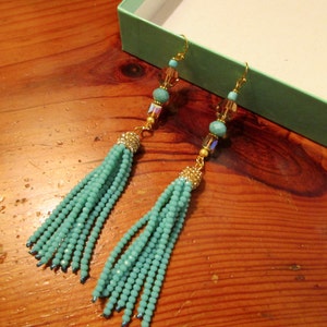 May include: A pair of turquoise beaded tassel earrings with gold-colored accents. The earrings feature small turquoise beads, gold-colored hooks, and decorative gold-colored caps. The earrings are displayed on a wooden surface.