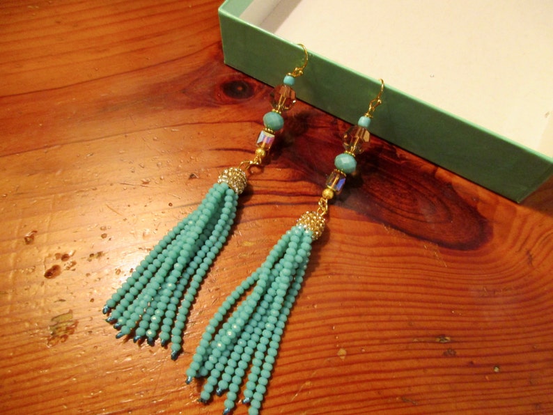 May include: A pair of turquoise beaded tassel earrings with gold-colored accents. The earrings feature small turquoise beads, gold-colored hooks, and decorative gold-colored caps. The earrings are displayed on a wooden surface.