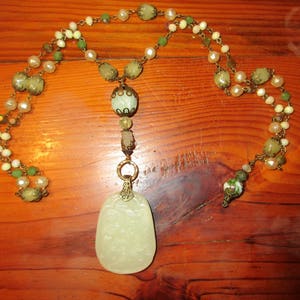 Puede incluir: Un collar largo con un colgante de jade. El collar est&aacute; hecho de metal dorado y tiene cuentas verdes, blancas y marrones.