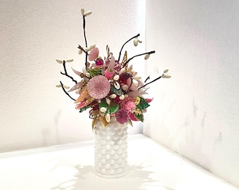 May include: A white ceramic vase with a textured surface holds a bouquet of pink, white, and green flowers made of plastic and glass. The flowers are arranged in a whimsical and artistic way.