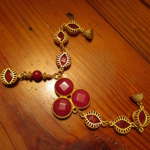 May include: A gold necklace with red gemstone accents. The necklace features a pendant with three large red gemstones and a smaller red gemstone pendant hanging from the bottom.