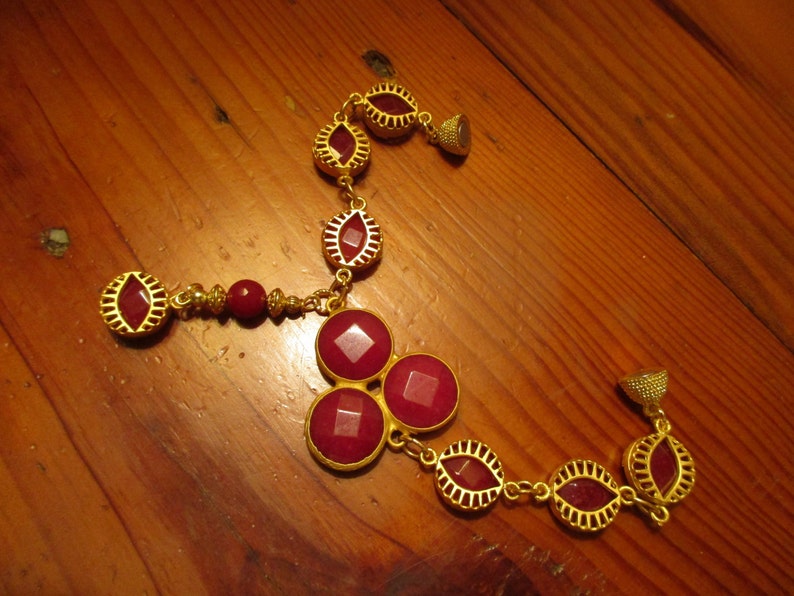 May include: A gold necklace with red gemstone accents. The necklace features a pendant with three large red gemstones and a smaller red gemstone pendant hanging from the bottom.