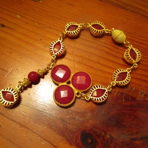 May include: A gold chain bracelet with a dangling charm featuring three red gemstones. The bracelet is adorned with gold oval-shaped links, each containing a red gemstone.