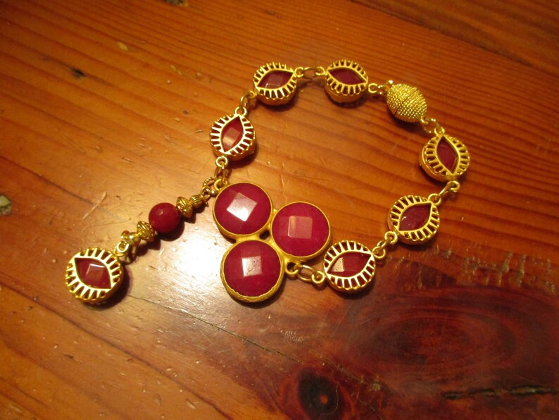 May include: A gold chain bracelet with a dangling charm featuring three red gemstones. The bracelet is adorned with gold oval-shaped links, each containing a red gemstone.