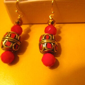 May include: A pair of gold earrings with red and gold beads. The beads are round and have a geometric pattern. The earrings are hanging from gold hooks.