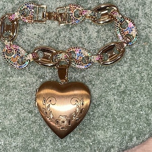 May include: A gold charm bracelet with a heart-shaped locket. The bracelet is made of gold chain links with colorful gemstones. The locket has a floral design.