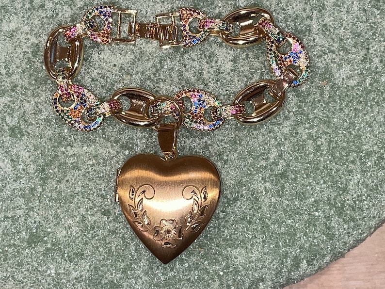 May include: A gold charm bracelet with a heart-shaped locket. The bracelet is made of gold chain links with colorful gemstones. The locket has a floral design.