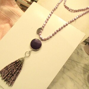 May include: A long, double-strand necklace made of light purple beads with a large, round, dark purple pendant and a tassel made of smaller purple beads.