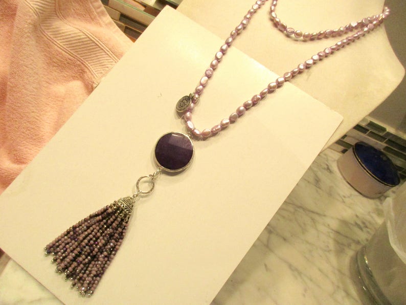 May include: A long, double-strand necklace made of light purple beads with a large, round, dark purple pendant and a tassel made of smaller purple beads.