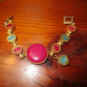Puede incluir: Un brazalete de oro con una piedra grande, redonda, de color fucsia rodeada de piedras más pequeñas, ovaladas, en tonos de verde azulado y rojo.