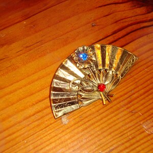 Puede incluir: Un broche de color dorado en forma de abanico con un dise&ntilde;o de pedrer&iacute;a azul y roja.