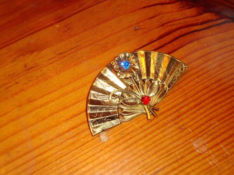 Puede incluir: Un broche de color dorado en forma de abanico con un dise&ntilde;o de pedrer&iacute;a azul y roja.