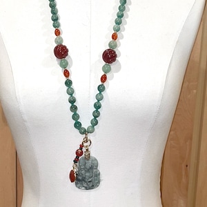 Puede incluir: Un collar largo de cuentas verdes y rojas con un colgante de jade tallado. El colgante tiene forma de persona y tiene un acento dorado.