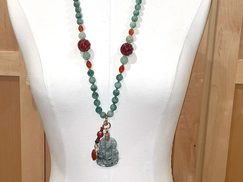 Puede incluir: Un collar largo de cuentas verdes y rojas con un colgante de jade tallado. El colgante tiene forma de persona y tiene un acento dorado.