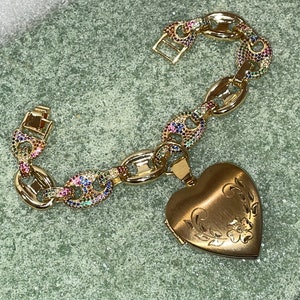 May include: A gold chain bracelet with a heart-shaped locket. The chain is decorated with colorful gemstones. The locket has an engraved floral design.