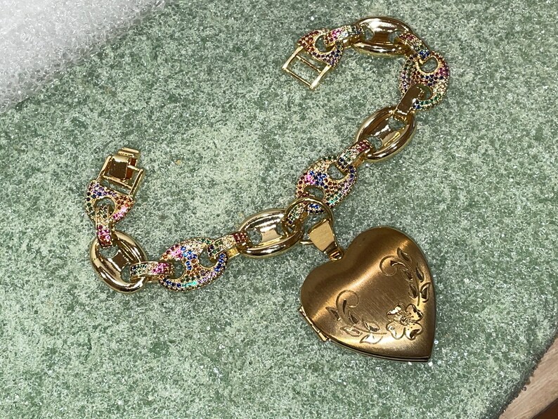May include: A gold chain bracelet with a heart-shaped locket. The chain is decorated with colorful gemstones. The locket has an engraved floral design.