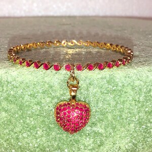May include: A gold bangle bracelet with a heart-shaped pendant. Both the bracelet and pendant are adorned with small, red gemstones.