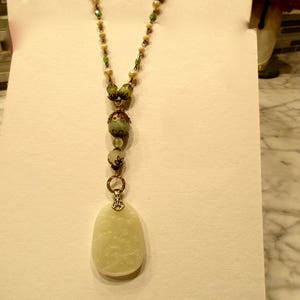 Puede incluir: Un collar largo de cuentas verdes y blancas con un gran colgante verde claro tallado.