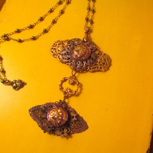 Może przedstawiać: Złoty łańcuszek z dwoma ozdobnymi wisiorkami. Wisiorki są wykonane z metalu z filigranowym wzorem i mają fioletowo-złote centrum. Łańcuszek leży na ż&oacute;łtym tle.