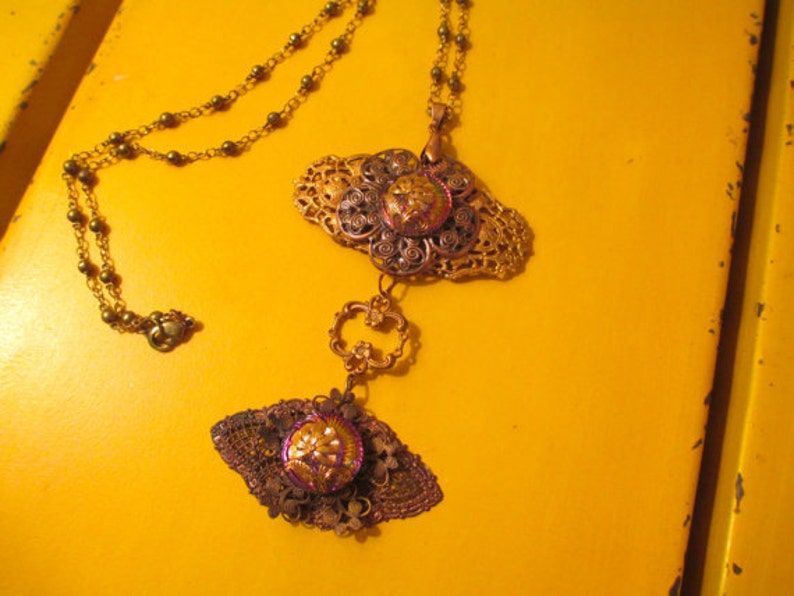 Może przedstawiać: Złoty łańcuszek z dwoma ozdobnymi wisiorkami. Wisiorki są wykonane z metalu z filigranowym wzorem i mają fioletowo-złote centrum. Łańcuszek leży na ż&oacute;łtym tle.