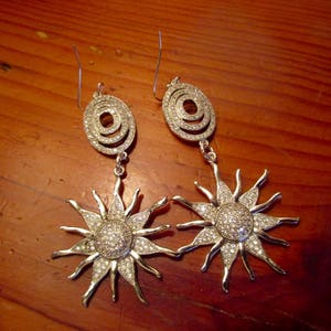 May include: A pair of silver earrings with a sunburst design. Each earring features a circular pendant with a smaller circle inside, adorned with clear rhinestones.