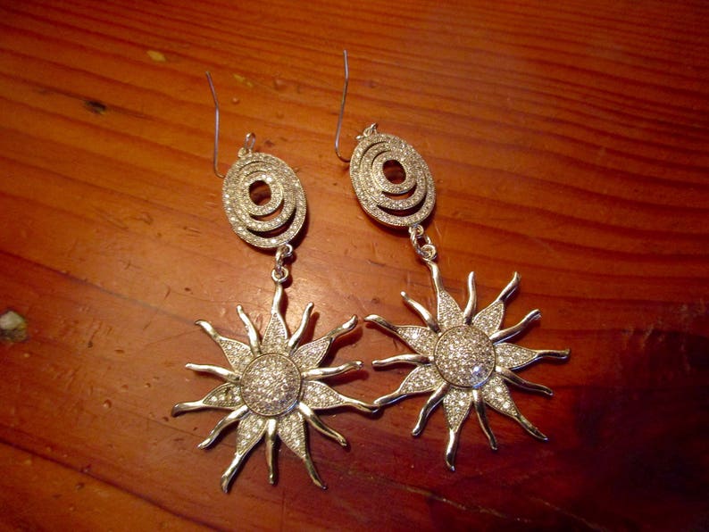 May include: A pair of silver earrings with a sunburst design. Each earring features a circular pendant with a smaller circle inside, adorned with clear rhinestones.