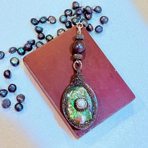 May include: Abalone shell pendant necklace with a pearl center and dark beaded accents. The pendant is set on a dark red rectangular surface, with a strand of dark pearl beads. The necklace is a unique piece of jewelry.