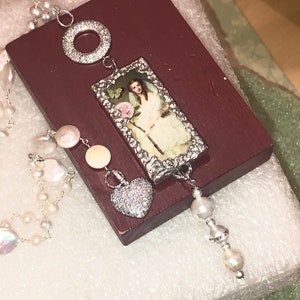 Pu&ograve; includere: Una collana d'argento con una perla bianca e un ciondolo a forma di cuore in argento. La collana presenta anche un ciondolo rettangolare con un'immagine vintage di una donna che tiene una rosa.