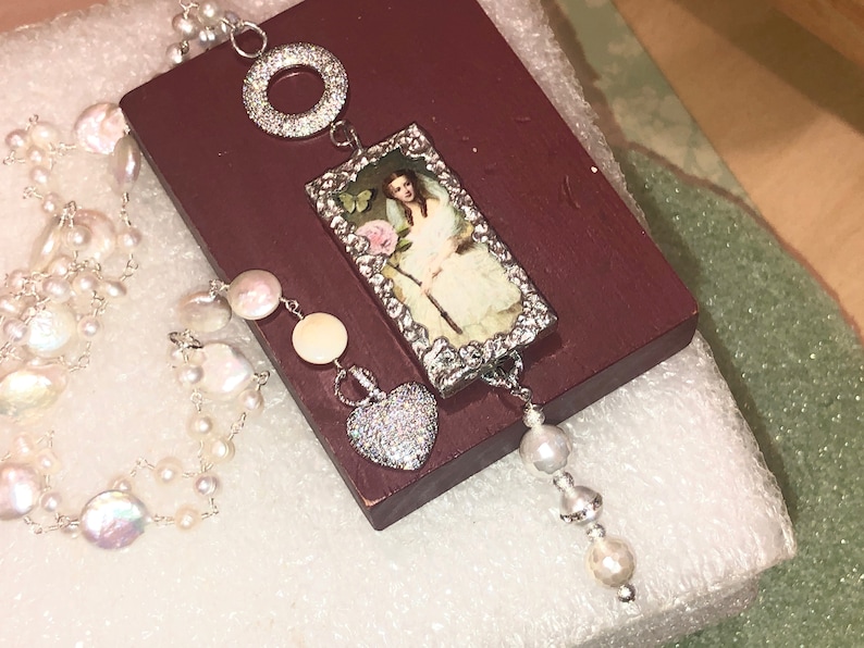 Pu&ograve; includere: Una collana d'argento con una perla bianca e un ciondolo a forma di cuore in argento. La collana presenta anche un ciondolo rettangolare con un'immagine vintage di una donna che tiene una rosa.