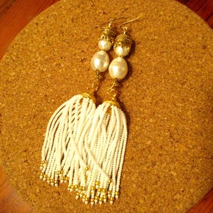 May include: A pair of white tassel earrings with gold accents and pearl-like beads. The earrings are hanging from a cork surface.