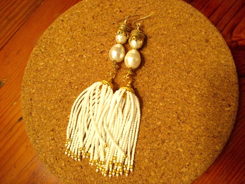 May include: A pair of white tassel earrings with gold accents and pearl-like beads. The earrings are hanging from a cork surface.