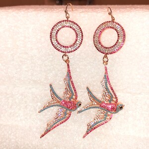 Puede incluir: Un par de pendientes colgantes con un dise&ntilde;o de p&aacute;jaro rosa y azul. Los pendientes presentan un c&iacute;rculo redondo, rosa y blanco incrustado de cristales.