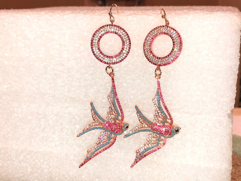 Puede incluir: Un par de pendientes colgantes con un dise&ntilde;o de p&aacute;jaro rosa y azul. Los pendientes presentan un c&iacute;rculo redondo, rosa y blanco incrustado de cristales.
