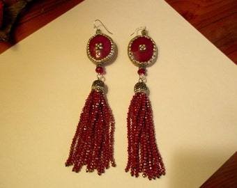 May include: A pair of red tassel earrings with a round, red, and gold-toned pendant. The earrings are made of beads and have a silver-toned hook.