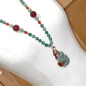 Puede incluir: Un collar de jade verde con cuentas rojas y naranjas y un colgante de jade. El colgante est&aacute; tallado con una figura y tiene un acento dorado.