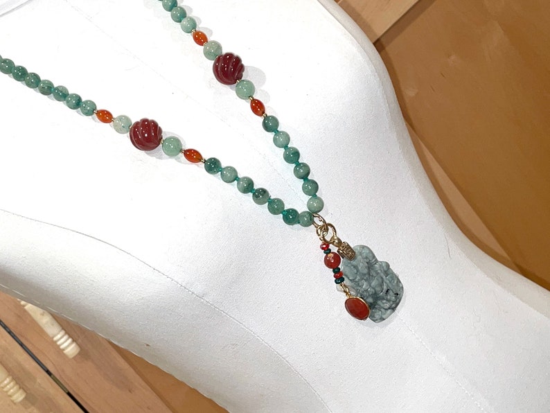 Puede incluir: Un collar de jade verde con cuentas rojas y naranjas y un colgante de jade. El colgante est&aacute; tallado con una figura y tiene un acento dorado.