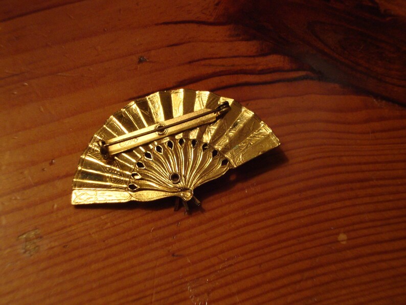 Puede incluir: Un broche de metal dorado en forma de abanico con un dise&ntilde;o decorativo. El broche tiene un pasador en la parte posterior para sujetarlo a la ropa.