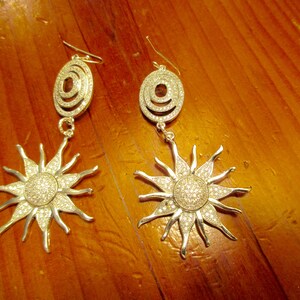 May include: A pair of silver earrings with a sunburst design. Each earring features a circular pendant with a smaller circle inside, and a larger sunburst design with sparkling rhinestones.