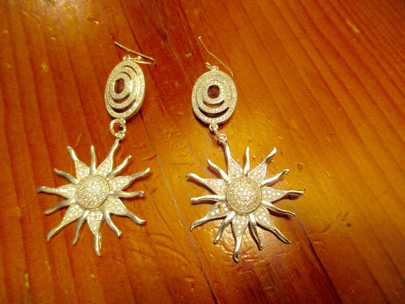 May include: A pair of silver earrings with a sunburst design. Each earring features a circular pendant with a smaller circle inside, and a larger sunburst design with sparkling rhinestones.