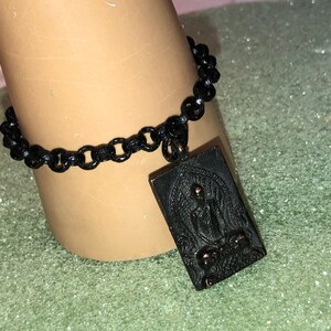May include: Black chain bracelet with a rectangular pendant featuring a detailed engraved image of a Buddha.
