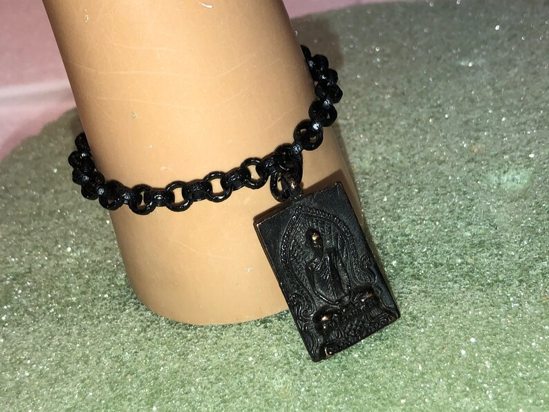 May include: Black chain bracelet with a rectangular pendant featuring a detailed engraved image of a Buddha.