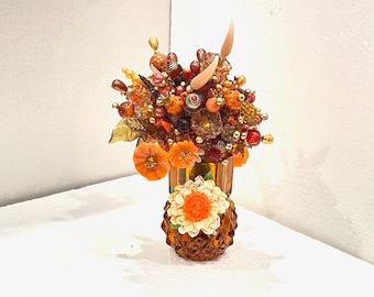 May include: A small, amber glass vase with a textured surface holds a bouquet of artificial flowers. The flowers are made of glass beads and feature orange, red, and gold colours. The vase is decorated with a single orange flower.