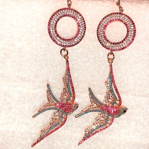 Puede incluir: Un par de pendientes de tono dorado con un dise&ntilde;o circular y un colgante de p&aacute;jaro golondrina. Los pendientes est&aacute;n adornados con cristales rosas y azules.