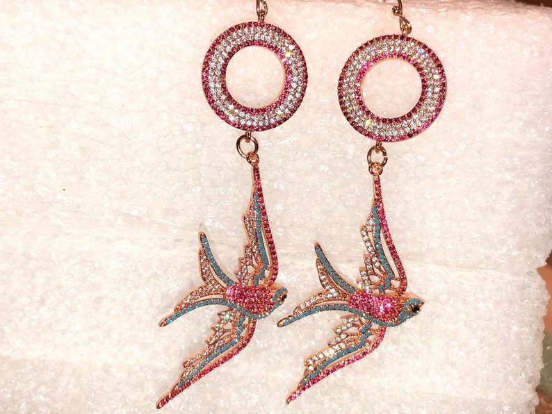 Puede incluir: Un par de pendientes de tono dorado con un dise&ntilde;o circular y un colgante de p&aacute;jaro golondrina. Los pendientes est&aacute;n adornados con cristales rosas y azules.