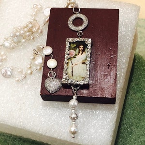 Pu&ograve; includere: Una collana di perle bianche con un ciondolo d'argento che presenta un'immagine incorniciata di una donna con un abito bianco che tiene un fiore rosa, un cerchio d'argento e un cuore d'argento con strass trasparenti.