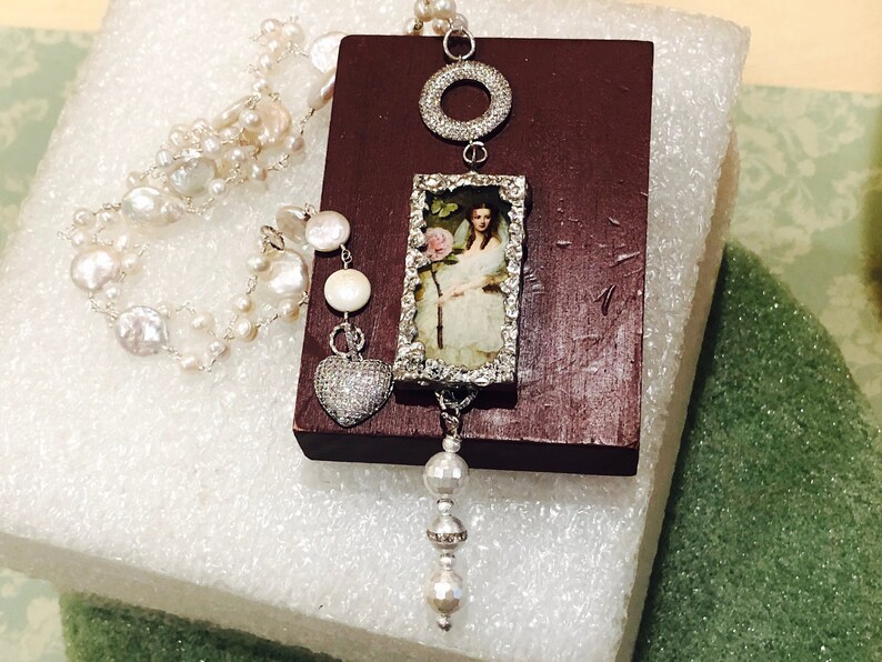 Pu&ograve; includere: Una collana di perle bianche con un ciondolo d'argento che presenta un'immagine incorniciata di una donna con un abito bianco che tiene un fiore rosa, un cerchio d'argento e un cuore d'argento con strass trasparenti.