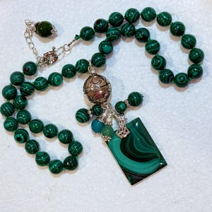 May include: A necklace featuring green malachite beads with dark green stripes. The necklace has a silver pendant with a rectangular malachite stone and a decorative silver charm. Additional green and blue beads are also present.