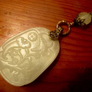Puede incluir: Un colgante de jade blanco con un dise&ntilde;o floral y un p&aacute;jaro, unido a una cadena dorada con un cierre decorativo.