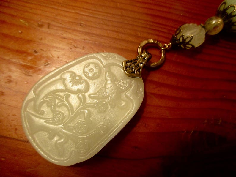Puede incluir: Un colgante de jade blanco con un dise&ntilde;o floral y un p&aacute;jaro, unido a una cadena dorada con un cierre decorativo.