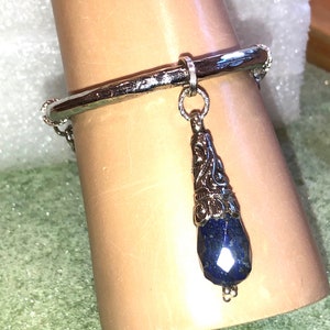 May include: A silver bangle bracelet with a dangling charm featuring a blue gemstone and intricate silver detailing.